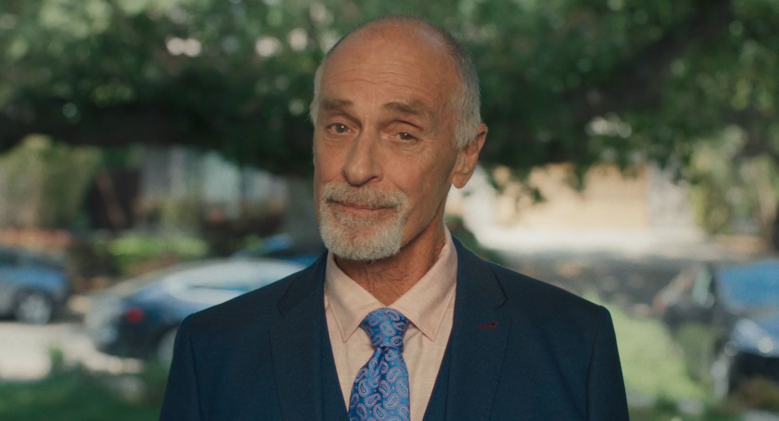 Keith Carradine as Dr. Wagner in a blue suit and patterned tie, smiling slightly.