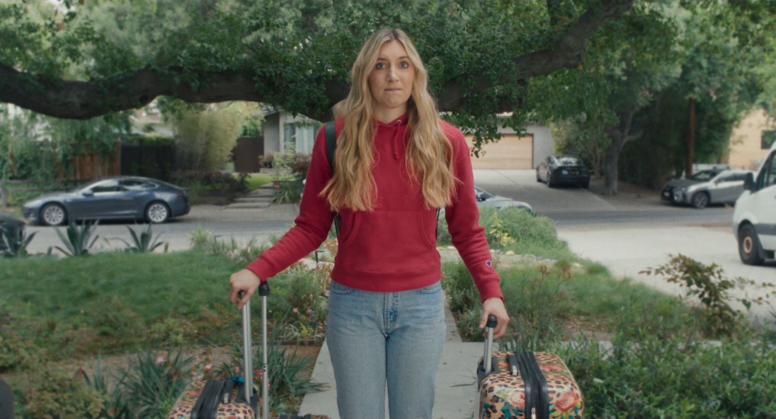 Lydia stands on a suburban sidewalk in a red hoodie with two rolling suitcases.
