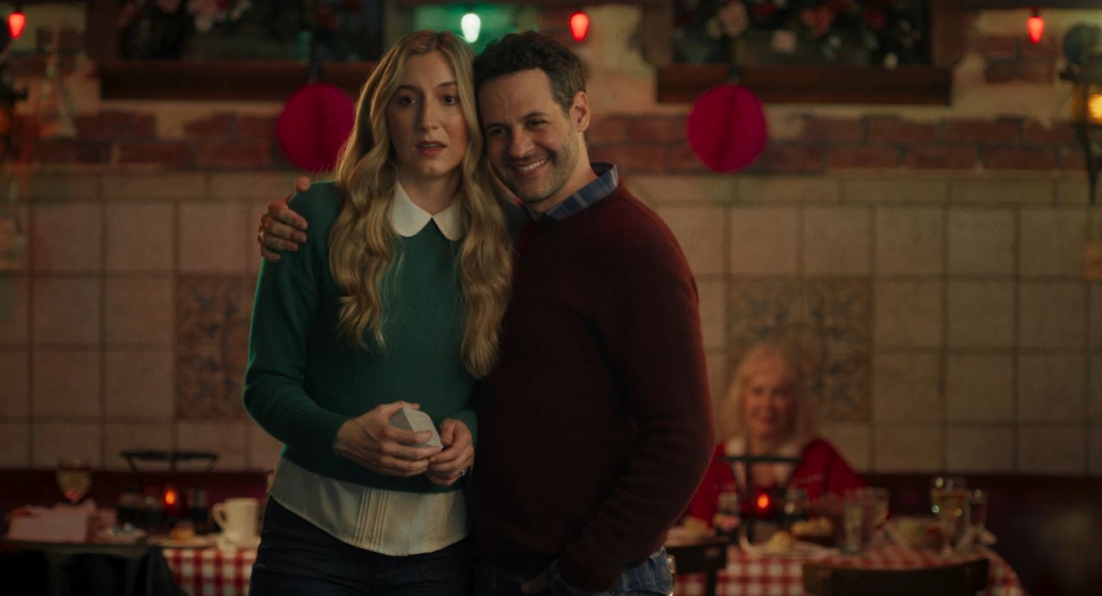 Lydia and Tim embrace in a restaurant decorated with red Christmas ornaments.